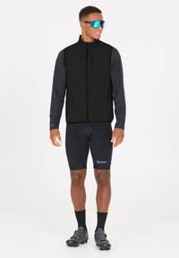 Svart ärmlös väst över en långärmad svart skjorta, tillsammans med svarta cykelbyxor och skor. Stora blå reflexglasögon.