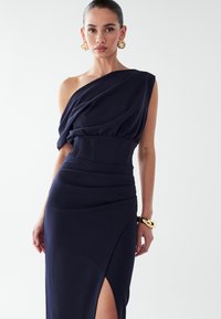 Vestido azul marino de un solo hombro con corsé ajustado y tela drapeada. Presenta una abertura lateral y una textura suave. Brazalete dorado en la muñeca.