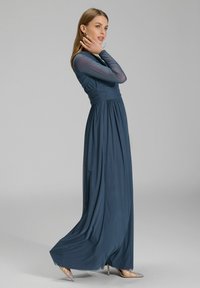 Pitkä, soljuva mekko syvän sinisenä, jossa on läpinäkyvät pitkät hihat ja vyötärö on kiristetty. Sileä tekstiili elegantilla laskostusilmalla ja nilkkoihin ulottuva malli.