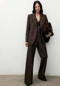 Completo in misto lana marrone con un blazer su misura, pantaloni a gamba larga e un gilet con bottoni. Gli accessori includono una borsa marrone soffice.