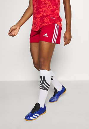 Persoon draagt rode Adidas sportshorts, witte kniekousen met zwarte patronen en blauwe zaalvoetbalschoenen, lopend.