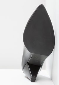 Chaussure à talon aigu en cuir noir, à bout pointu, inclinée vers le haut, montrant une semelle lisse et un talon aiguille sur un fond blanc.