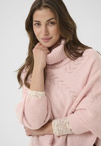 Ženska z dolgimi rjavimi lasmi, oblečena v roza pleteni pulover in kremno čipkastimi manšeti, naslonjena na dlan, se nežno smehlja pred enostavno ozadjem.