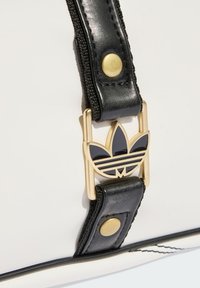 White bag with black accents, featuring a gold buckle and a black Adidas logo. Smooth texture with contrasting materials and stitching details.