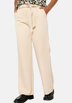 Beige wide-leg pants with a tied waistband, smooth texture, and side pockets. Worn with black patent shoes for contrast.