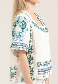 Blusa bianca in cotone con intricati motivi floreali blu e verdi e orlo a scallop. Presenta un collo quadrato e maniche ampie.