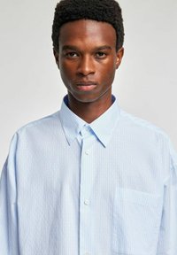Jasnoniebieska koszula w paski z kołnierzykiem na guziki i pojedynczą kieszenią na piersi; wykonana z miękkiego materiału o luźnym kroju.