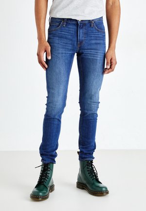 Blå denimjeans med smal passform, femfickdesign och blekta detaljer, i kombination med gröna snörskor och en grå T-shirt.