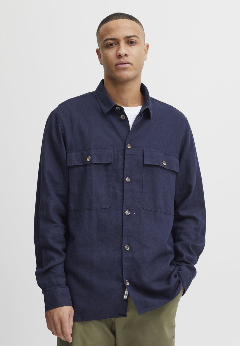 Camisa de botones azul marino hecha de tejido texturizado, con dos bolsillos en el pecho, botones marrones y mangas largas, combinada con pantalones de color claro.