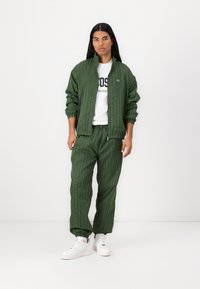 Grüner, texturierter Jogginganzug mit gestepptem Design, ausgestattet mit elastischen Bündchen und Bund, kombiniert mit einem weißen Hemd und weißen Sneakers.