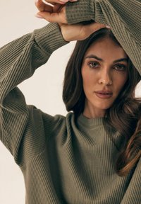 Sweter w kolorze oliwkowym, z żebrowanym wzorem i teksturowanym wykończeniem. Luźne, długie rękawy oraz okrągły dekolt. Ręce modelki uniesione, eksponujące detale rękawów.