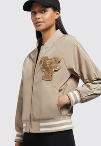 Beige bomberjack met een opvallende bruine tijgerpatch, gestreepte manchetten en een soepele textuur. Voorzien van een rits en zijzakken.
