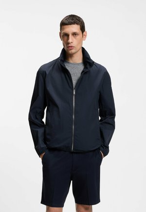 Junger Mann trägt eine marineblaue Jacke mit Reißverschluss über einem grauen Hemd und marineblauen Shorts und steht mit den Händen in den Taschen vor einem schlichten weißen Hintergrund.