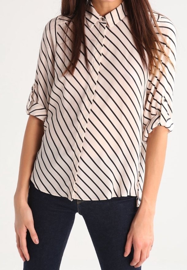 Femme portant une chemise blanche avec des rayures noires diagonales et des manches retroussées, associée à un jean bleu foncé.