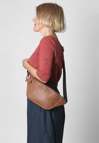 Borsa a tracolla in pelle marrone con superficie liscia, chiusura con zip e tracolla regolabile, indossata contro una gonna scura di lato.