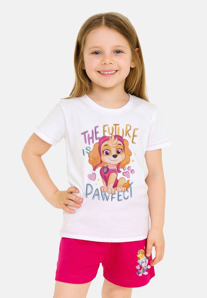Una giovane ragazza sorridente indossa una maglietta bianca con un cane dei cartoni animati e la scritta "Il futuro è perfetto", abbinata a pantaloni corti rosa brillante con personaggi di cani.