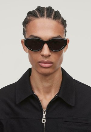 Black oval sunglasses with thick frames, featuring dark lenses. The model wears a black jacket with a zippered collar. Smooth texture.