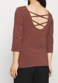 Brun långärmad topp med en djup ryggöppning och ett crisscross-stil bärande band. Mjuk tyg med en vanlig passform och fållavslutning.