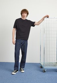 Black cotton t-shirt, relaxed fit, paired with dark denim pants featuring visible stitching. White sneakers, metal wire cart in background.