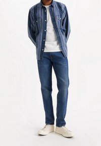 Blå denimskjorta med ljusa vertikala ränder över en vit t-shirt, matchad med blå jeans och beige sneakers. Avslappnad, lager-på-lager look.