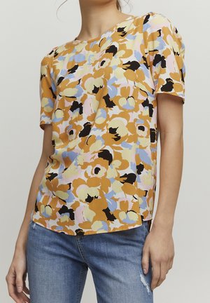 Woman wearing a short-sleeve floral top in mustard, black, blue, and pink paired with blue denim jeans against a plain background.