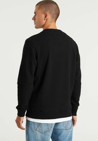 Schwarzes Langarm-Sweatshirt mit geripptem Muster, Rundhalsausschnitt und lässiger Passform; getragen über einem weißen Lagenshirt, kombiniert mit hellblauen Jeans.