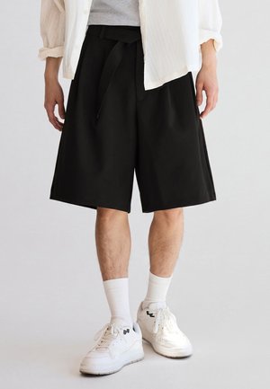 Man draagt zwarte knielange shorts, witte sneakers, witte sokken, een grijs shirt en een wit open overhemd, staand op een effen lichte achtergrond.