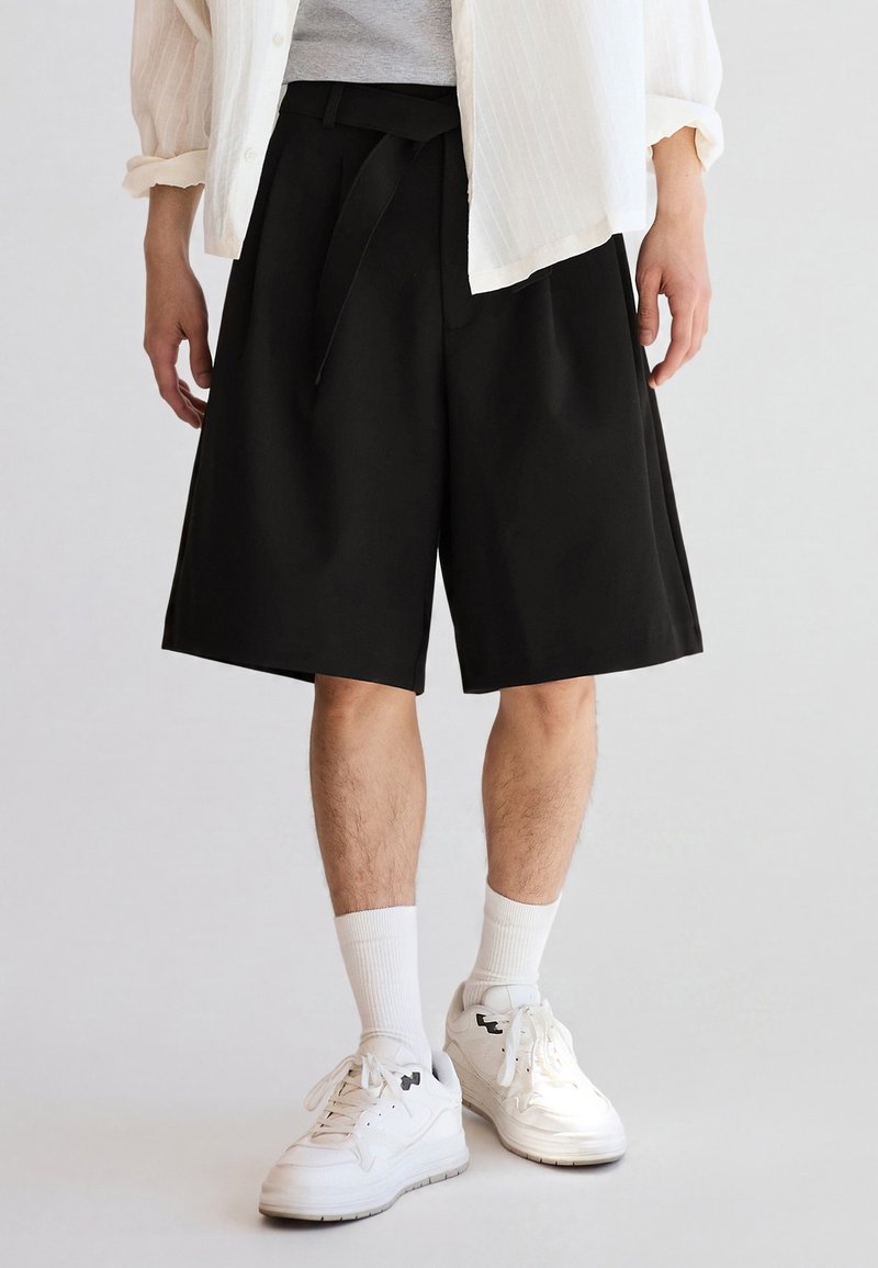 Man wearing black knee-length shorts, white sneakers, white socks, gray shirt, and white open button-down shirt standing on plain light background.