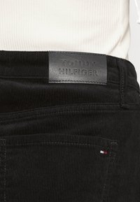 Svarta kordflorsjeans med en präglad läderetikett "TOMMY HILFIGER" på midjebandet. Har en liten röd, vit och blå accent på fickan.
