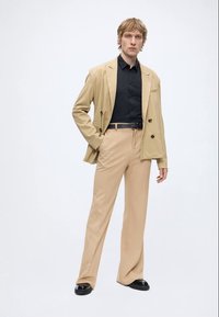 Ung mann som står med hånden i lommen, iført beige dress, svart skjorte, svart belte og svarte sko mot hvit bakgrunn.