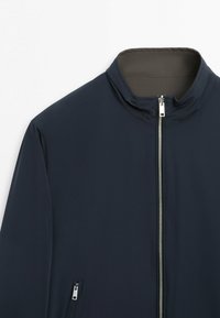 Marineblaue Jacke mit silbernem Frontreißverschluss und Reißverschlusstasche an der Seite, ausgestattet mit Stehkragen und glatter Stoffstruktur.