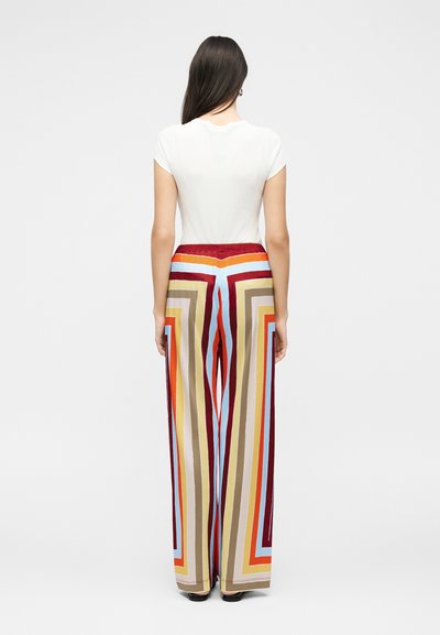 Γυναίκα με μακριά σκούρα μαλλιά φοράει άσπρο κοντομάνικο τοπ και ψηλόμεσο φαρδύ παντελόνι με πολύχρωμες γεωμετρικές ρίγες, όρθια με την πλάτη γυρισμένη.