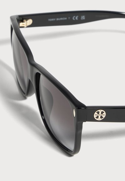 Gros plan de lunettes de soleil noires aux verres gris foncé, ornées d’emblèmes circulaires dorés sur les branches et posées sur une surface blanche.