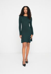Femme se tenant contre un fond blanc, portant une robe en tricot ajustée à manches longues de couleur vert foncé et des escarpins noirs à talons pointus.