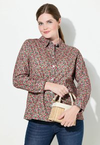 Camicia a fiori con bottoni in rosa e verde, con colletto a punta e maniche lunghe. Indossata con jeans scuri e tiene in mano una piccola borsa intrecciata.