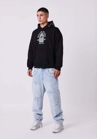 Felpa nera con stampa grafica, jeans denim leggero azzurro larghi con ricamo a forma di stella, e scarpe sportive bianche.