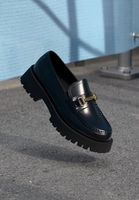 Juodas odinis loaferis su blizgiu paviršiumi, auksiniais detalėmis ir masyviu juodu padu. Savybėms priklauso klasikinis dizainas su matomomis siūlėmis.