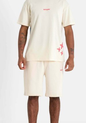 Ensemble en coton beige comprenant un t-shirt et un short avec un petit logo rouge sur la poitrine et des accents en forme d'étoile rose sur le short. Texture douce.