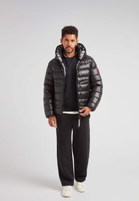 Giacca a vento nera con zip anteriore e cappuccio, dotata di cordini bianchi con logo e un'etichetta rossa sulla manica; abbinata a pantaloni scuri.