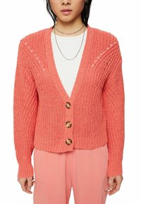 Koraalkleurige gebreide cardigan met v-hals, drie grote bruine knopen, decoratieve gaatjes, en ribbels aan de mouwen en onderkant. Gestyled over een witte top.