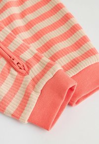 Lähivaade soonikkoelastsete varrukate kohta, mis on korall- ja beežis horisontaalsete triipudega ning millel on korallvärviline mansett ning ühe varruka korallvärvilise tõmblukuga detail.