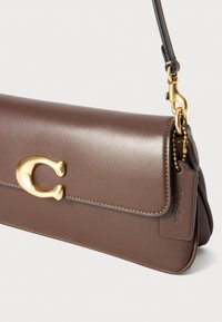 Braune Lederhandtasche mit glatter Textur, ausgestattet mit einem goldfarbigen Verschluss und einem abnehmbaren Tragegurt. Enthält ein Logoschild an der Seite.