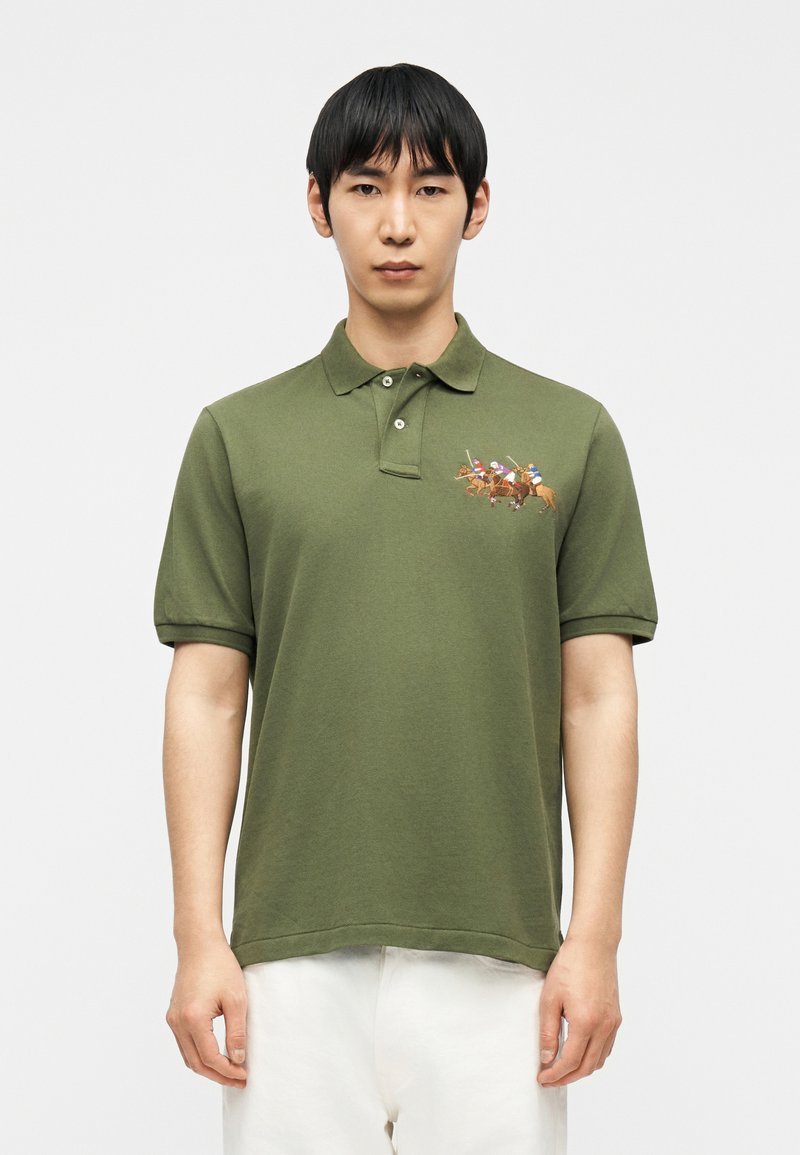 Άνδρας που φοράει πράσινο ελιάς πόλο πουκάμισο με κεντητό σχέδιο τριών παίκτων πόλο πάνω σε άλογα, συνδυασμένο με λευκό παντελόνι, κοιτάζοντας μπροστά.