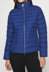 Sininen puhallettava takki, jossa on korkea kaulus, tilkkutekstuuri, etuvetoketju ja pieni logo rinnassa. Sopii kylmille säille.