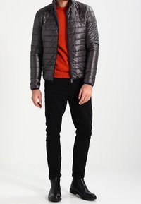 Dunkelgraue Daunenjacke mit gestepptem Design, leichtem Material und hohem Kragen, kombiniert mit einem leuchtend orangefarbenen Pullover und schwarzen Hosen.