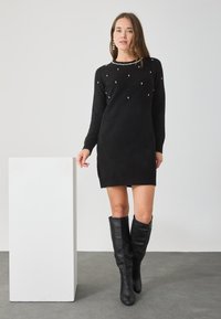 Fekete kötött ruha kerek nyakkivágással és szétszórt díszítésekkel. Térd fölé érő fekete bőr csizmával viselve. Egyszerű, testre simuló fazon.