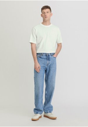 Witte t-shirt met korte mouwen, gecombineerd met lichtblauwe straight-leg jeans. Model staat met één hand in de zak en draagt witte sneakers.