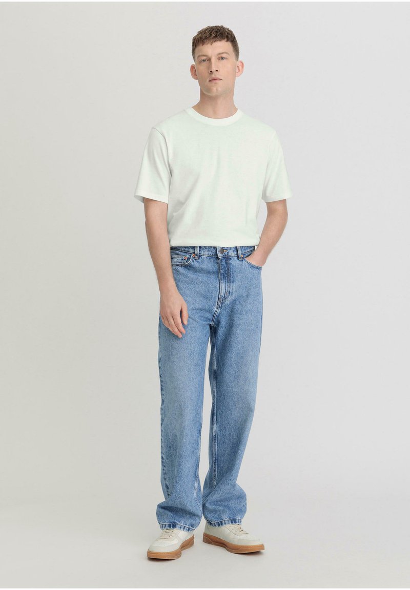 Weißes T-Shirt mit kurzen Ärmeln, kombiniert mit hellblauen geraden Jeans. Das Modell steht mit einer Hand in der Tasche und trägt weiße Sneakers.