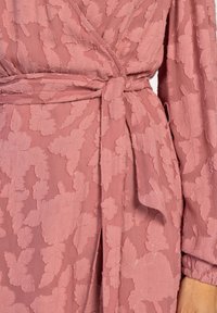Roze wikkeljurk van textuurstof, met bloemenborduring en een geknoopt riem bij de taille. Lange mouwen met geplooide details.