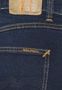 Jeans de denim azul escuro com costura amarela em contraste e um rótulo de couro no cós com gráficos laranja. Pequeno bolso com detalhe do logotipo.